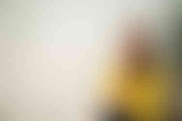 Gold sequins reflecting on white wall like disco ball with therapist in yellow shirt smiling at the camera, sat in a rattan peacock chair.