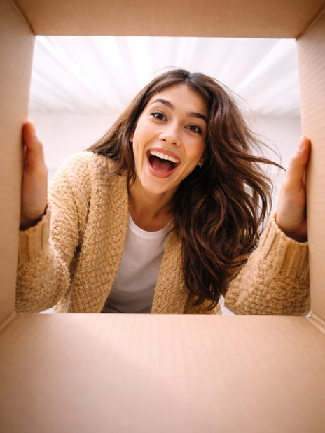 A smiling woman with long hair looks into a box from the outside, her face framed by the cardboard flaps, creating a playful perspective.