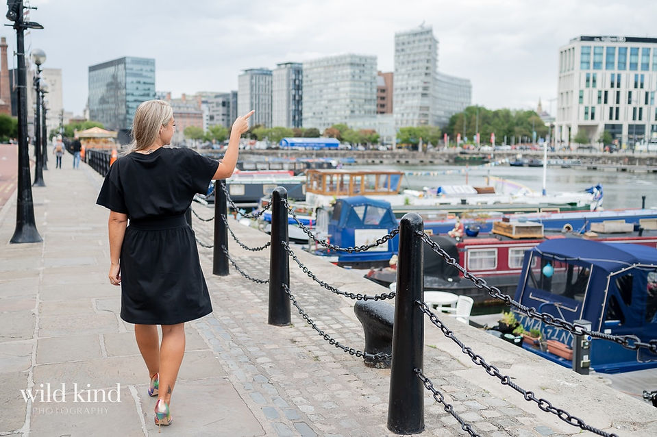 Vicki Peers walking along Albert Docks pointing at Liverpool skyline.