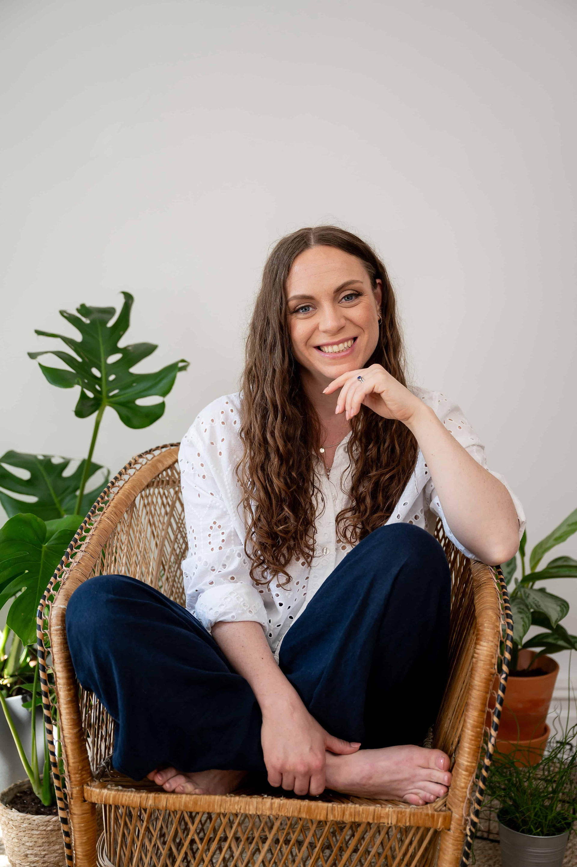A person with long hair, wearing a white shirt and dark pants, sits barefoot on a wicker chair. Green plants are in the background.