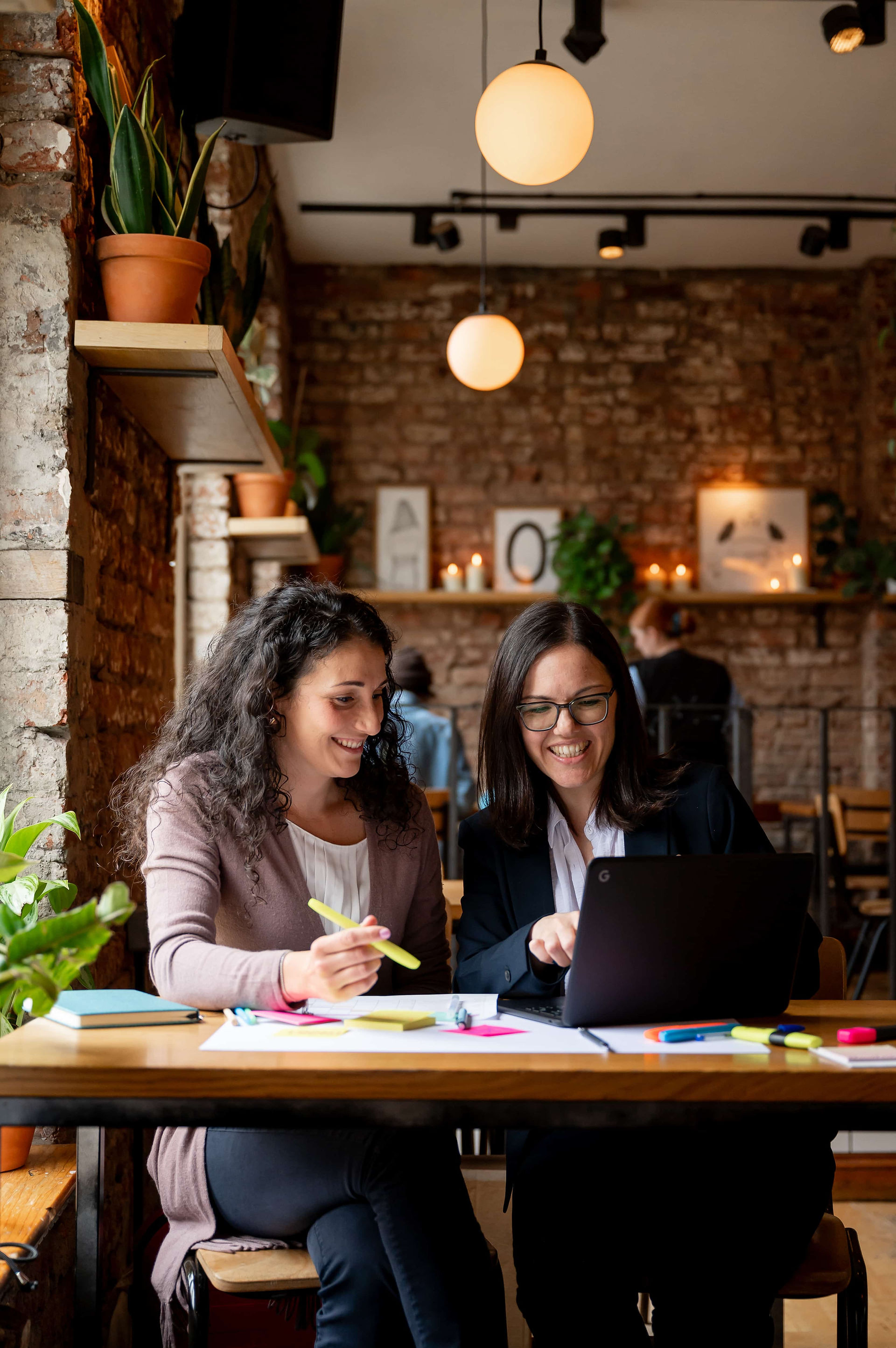 Two women smiling and working together on a laptop in a cozy café decorated with plants and hanging lights during a Liverpool branding photoshoot.