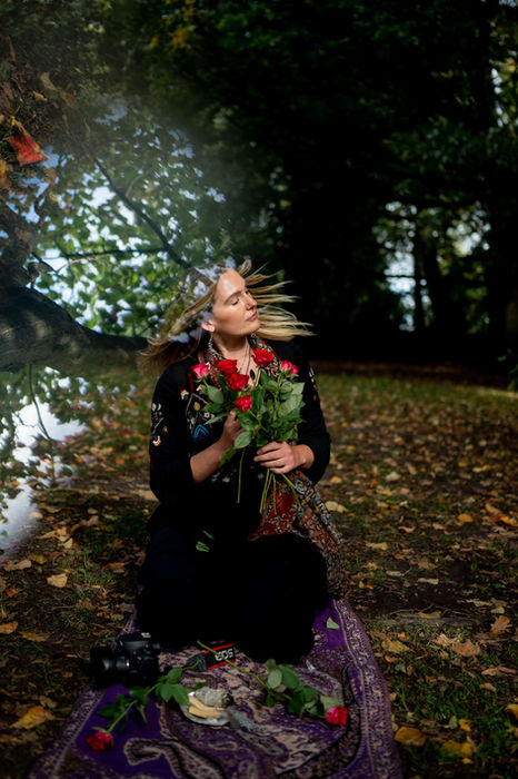 A woman in traditional attire sitting on a patterned blanket outdoors, holding roses and surrounded by autumn leaves for a brand photoshoot.