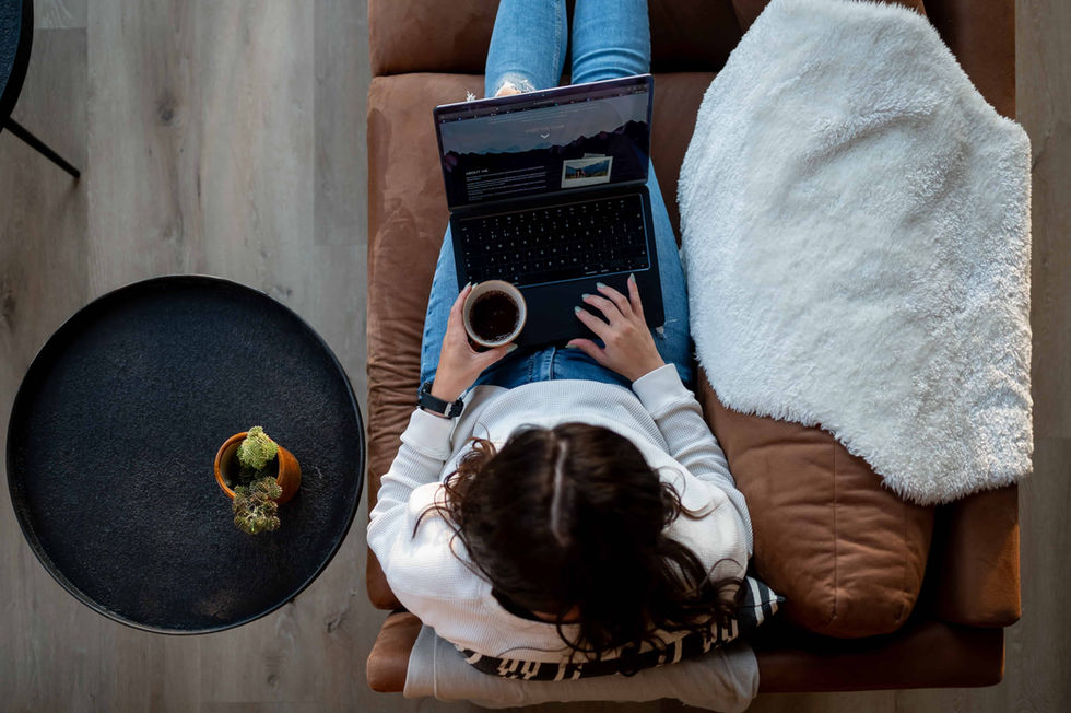 Over head shot of the lady with her legs stretched out on brown sofa. open laptop resting on her legs and cup in her left hand. 
