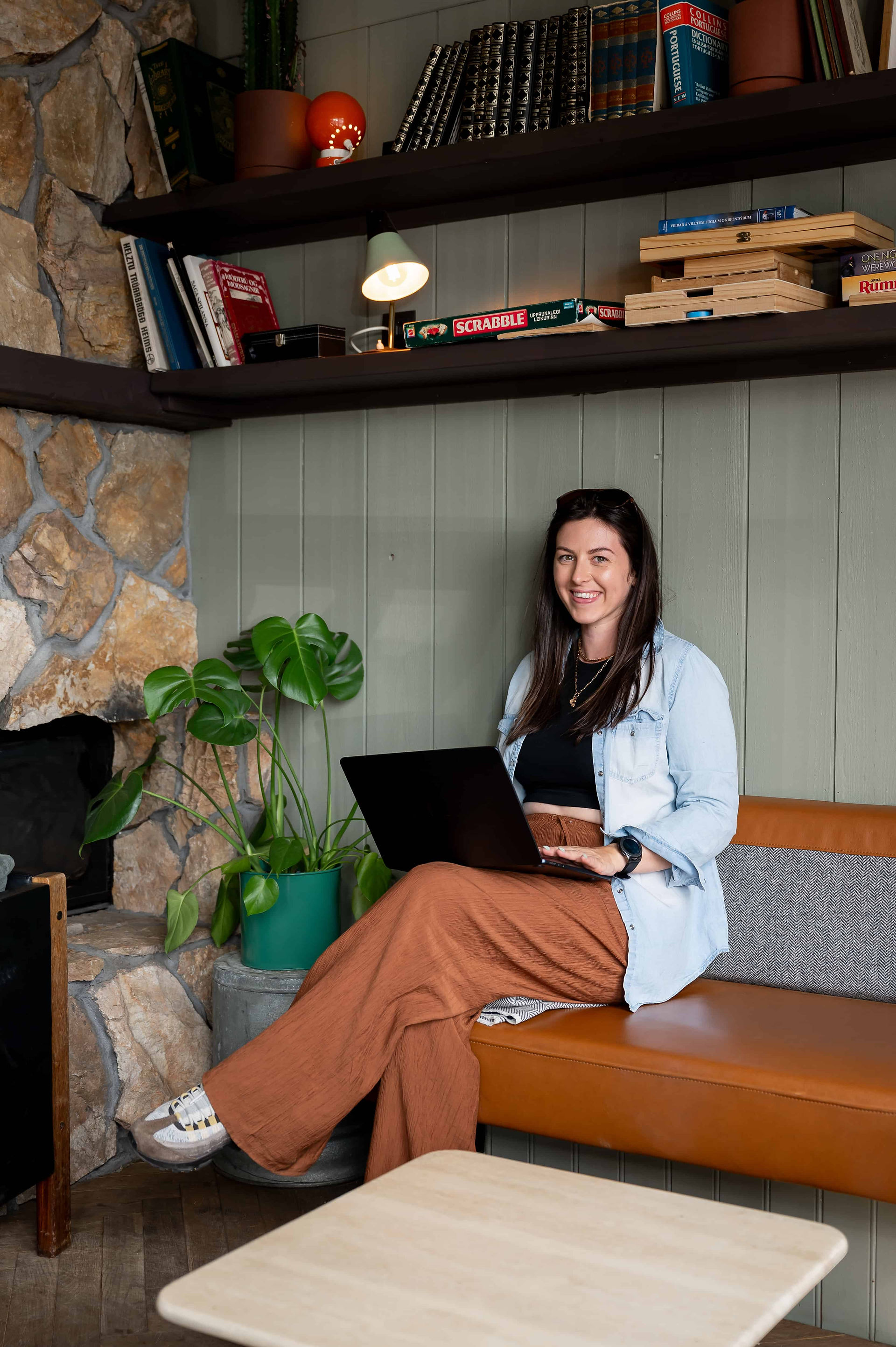 Lady sat on bench inside, her legs are crossed and a laptop in resting on her top leg. there is 2 long book shelves above her head on the wall.