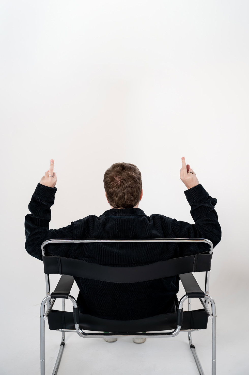 Back view of one founder with middle fingers up on both hands playfully while seated in a director’s chair during the studio photography session.