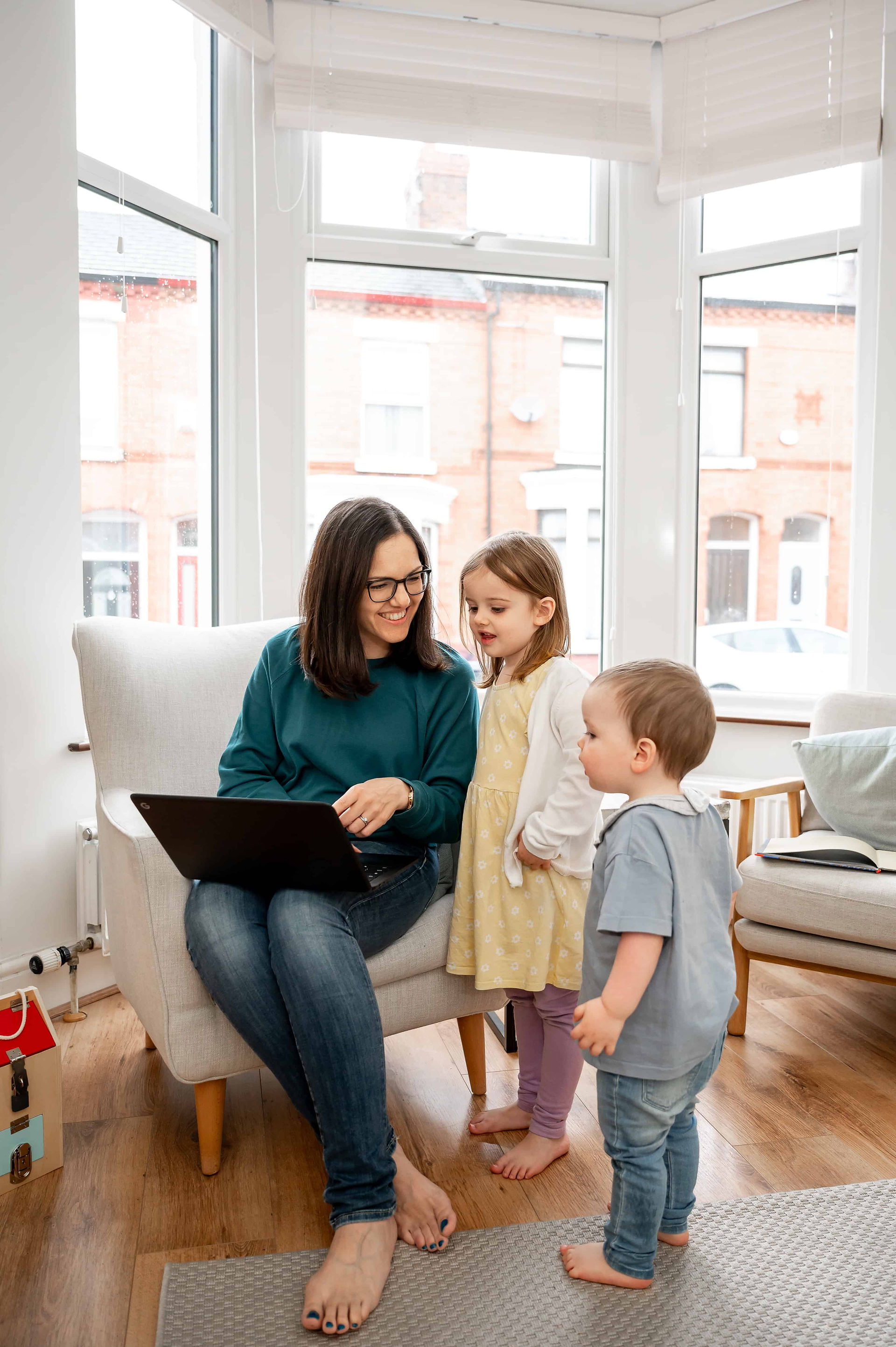 A woman sitting in a chair using a laptop, smiling at two young children standing beside her in a bright room with large windows during a Liverpool branding photoshoot.