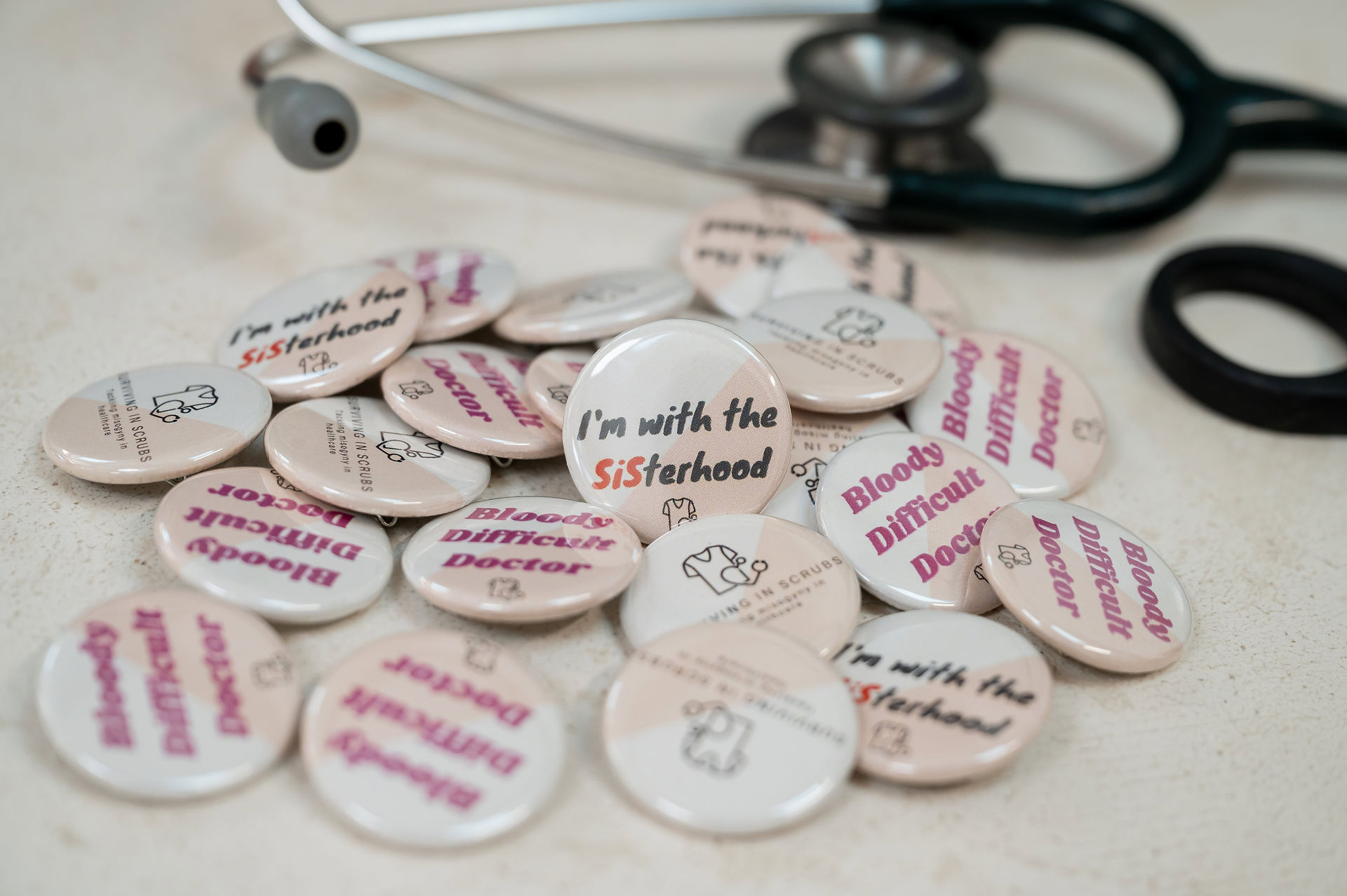 A pile of pins with phrases like "I’m with the Sisterhood" and "Bloody Difficult Doctor" lie next to a stethoscope on a table, capturing the essence of surviving in scrubs during a brand shoot.