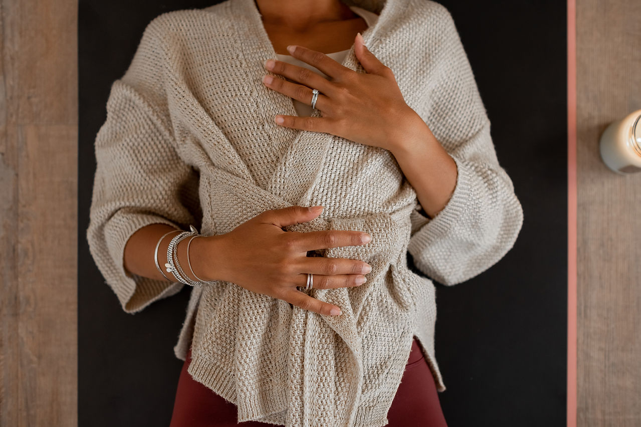 Person in a beige cardigan with hands placed on their chest and abdomen lying down on a dark surface, next to a lit candle, captured elegantly as part of a personal brand photographer's portfolio.