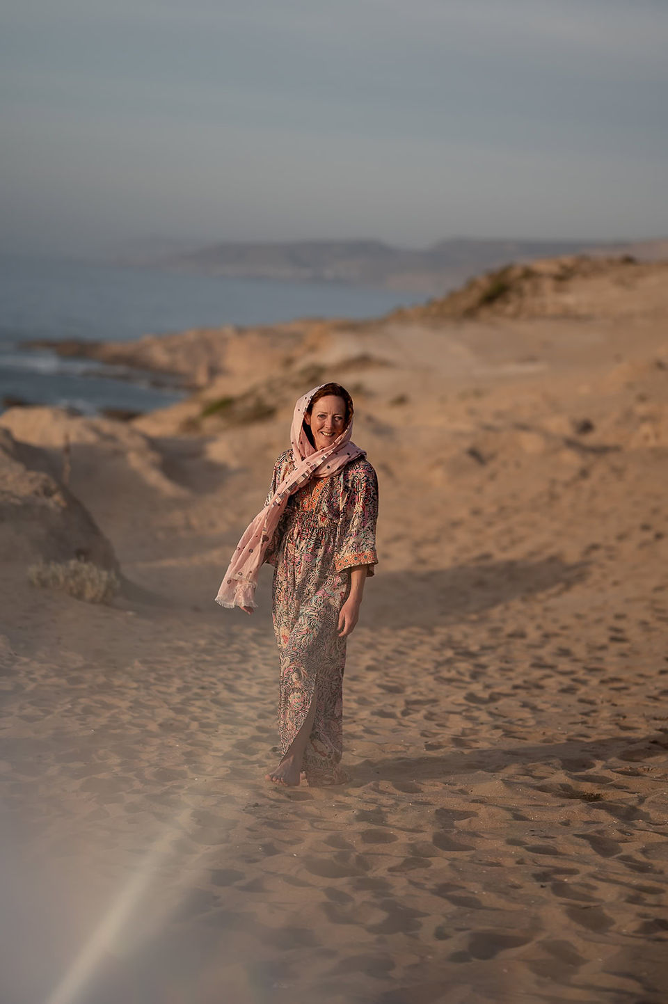 A person in a patterned dress and scarf walks along a sandy beach with distant hills and water visible under a clear sky.