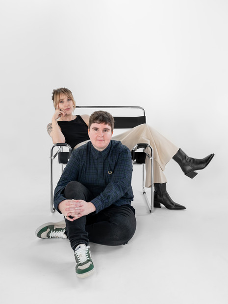Two business partners posing together during a brand photoshoot, one seated in a chair and one on the floor, editorial style studio shot.