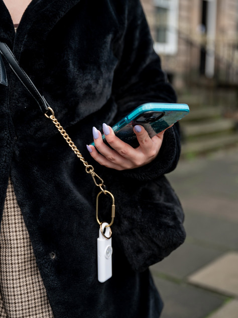 Close-up of a UK-legal personal safety alarm attached to a bag while the user holds a phone outdoors, photographed for Get Personal With Your Safety.