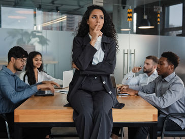 Businesswoman in a pinstripe suit sits on a table, pondering, as four colleagues work at laptops in a modern office. Mood is focused.