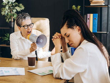 Person with megaphone yelling at another with closed eyes, hands clasped. Office setting, papers, coffee cup; tense mood, sunlight.
