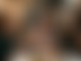 Five people sit around a table with drinks, one laughing, others looking displeased. Brick wall and hanging lights create a cozy setting.