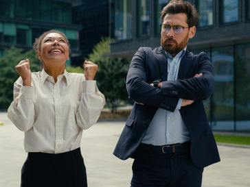 Woman in white shirt jubilantly raises fists beside a serious man in a suit, standing outdoors near modern glass buildings; trees in background.