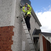 Technician installs satellite dish on house with ladder, blue sky, Munster Satellite Services.