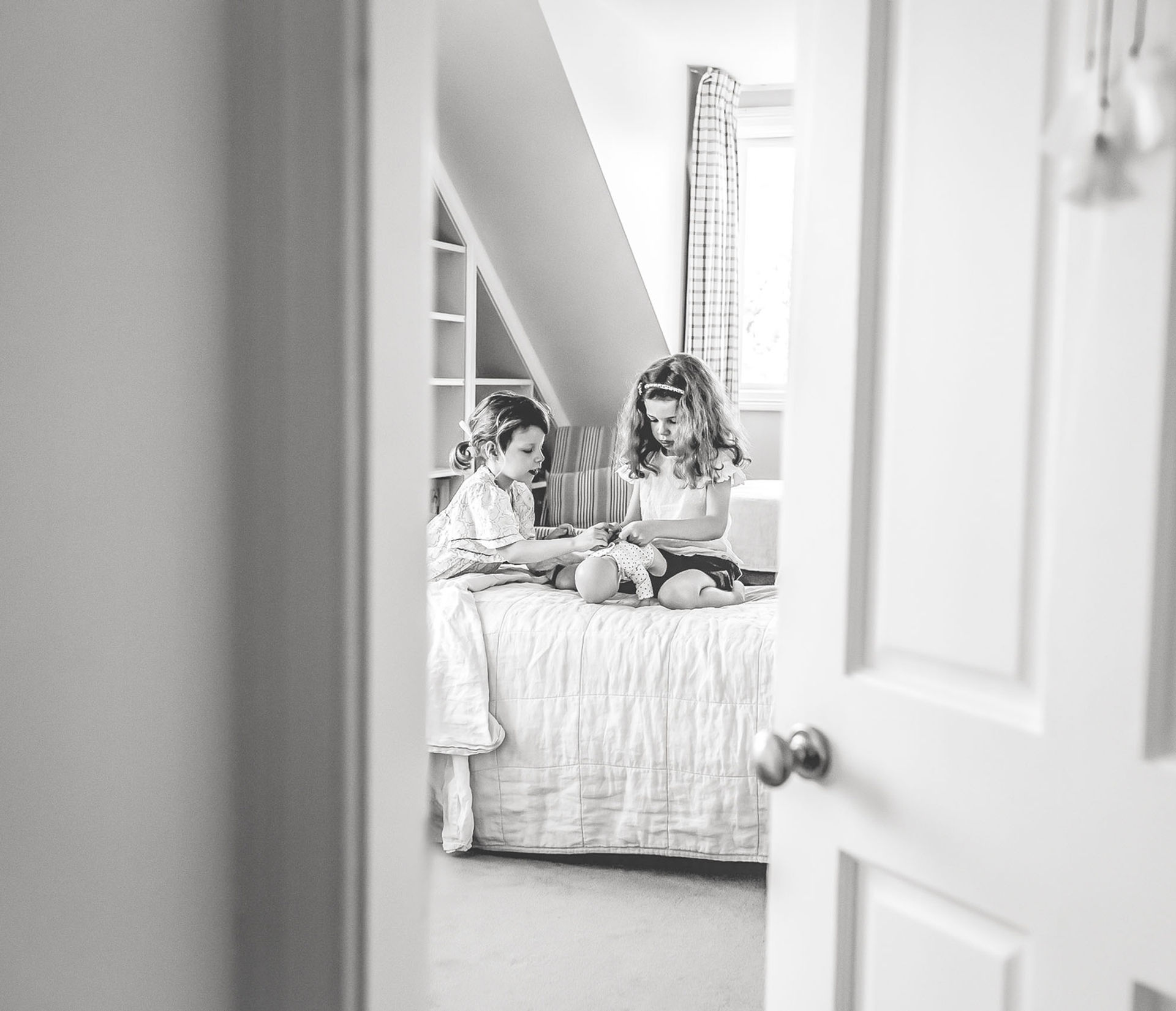 Candid shot in black and white of two sisters playing with their dolls looking through their door. Family Photographer Suzanne Stevenson Mosman Sydney