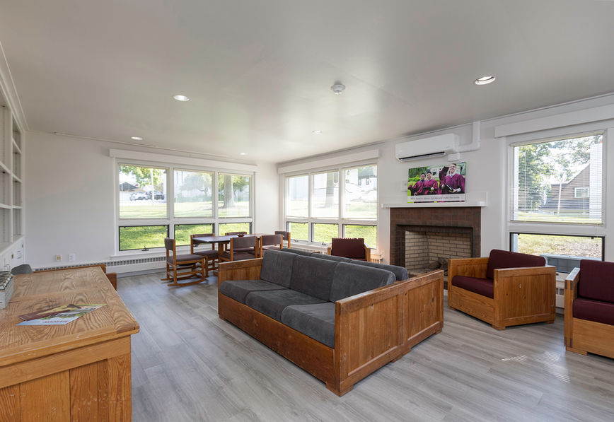 Bright room with wooden furniture, gray sofa, and chairs. Large windows overlook a green lawn. Fireplace with a colorful photo above. Cozy atmosphere.