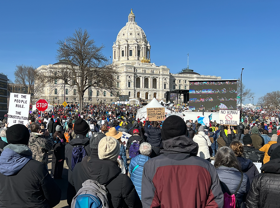 Crowd protesting with signs at a government building. Clear sky, some trees visible. Key signs read "We the People Rule", "No Human is Illegal", and "When injustice becomes law, resistance becomes duty."