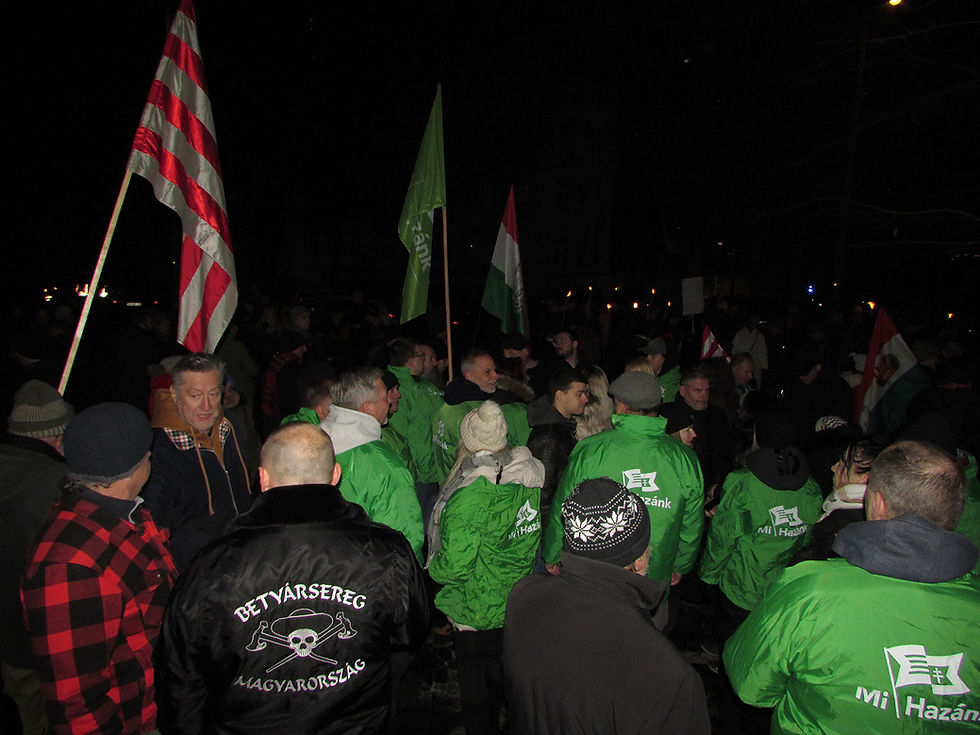 Nighttime crowd in green jackets holding Hungarian flags; black jacket reads BetyárSereg Magyarország, a neo-Nazi and far-right group.
