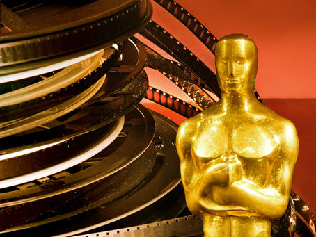 A golden statuette stands beside stacked film reels against a warm orange backdrop. The shiny surfaces and classic elements suggest a cinematic theme.