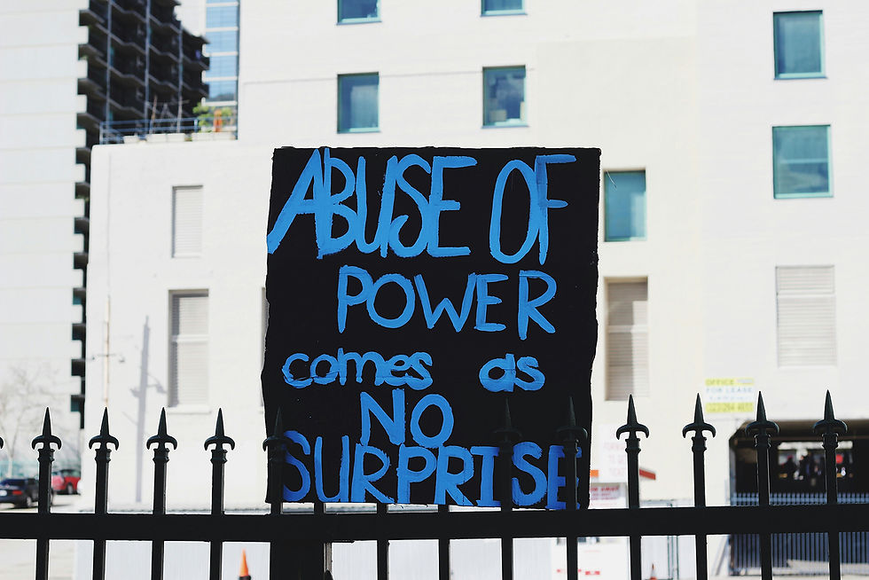 Sign with blue text "ABUSE OF POWER comes as NO SURPRISE" on black background mounted on a fence, with buildings in the background.