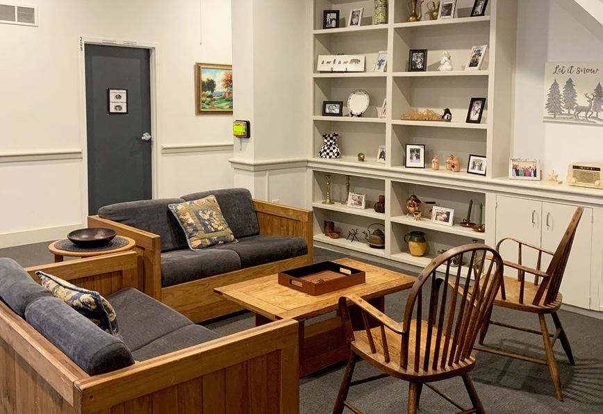 Cozy room with wooden furniture, gray couches, patterned pillows, bookshelves with photos, art on walls, and a gray door. Comfortable ambiance.