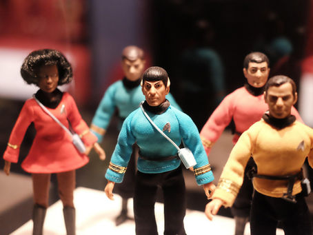 Action figures dressed in Star Trek uniforms stand together. Blue, red, and yellow outfits are visible, with a blurry, dark background.