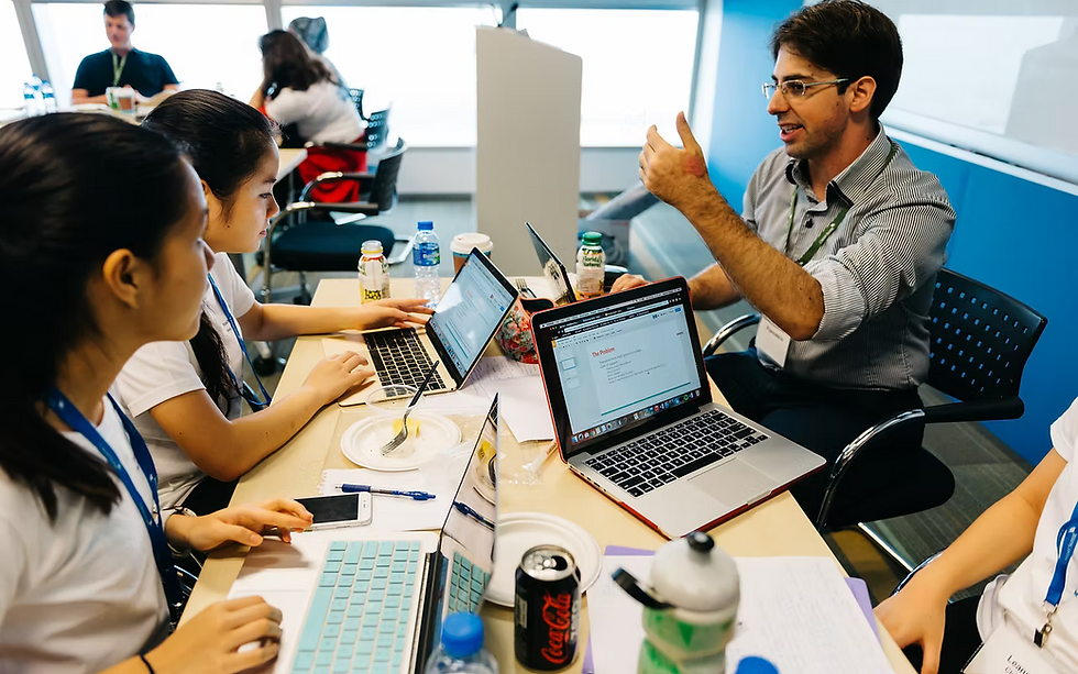 Man instructor teaches young women coding on laptops in classroom setting.