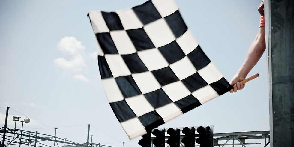 A hand waves a black and white checkered flag at a race finish line against a clear blue sky. Speaker poles are visible in the background.