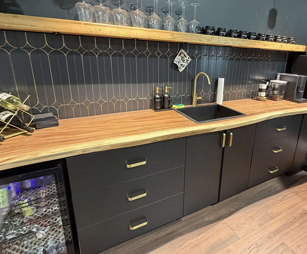 Stylish modern office coffee bar with black cabinets, live edge wood counter, brass sink, glasses, bottles, and a dark patterned backsplash