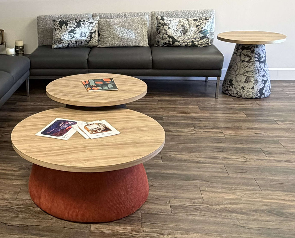 Modern lounge with graysofa and floral pillows, round wood and upholstered base coffee tables, and brochures on a wood floor.