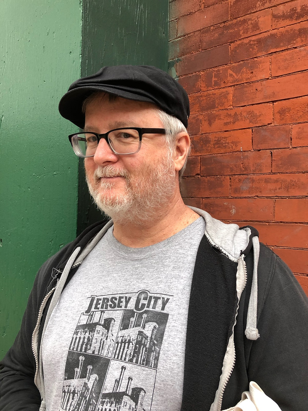 Jack is shown standing against a wall, part red brick & part wood painted 2 shades of green. He has a grey beard & hair. Is wearing black chunky glasses, black cap, a black unzipped hoodie, and a grey Jersey City t-shirt with 4 images of The Powerhouse Building.