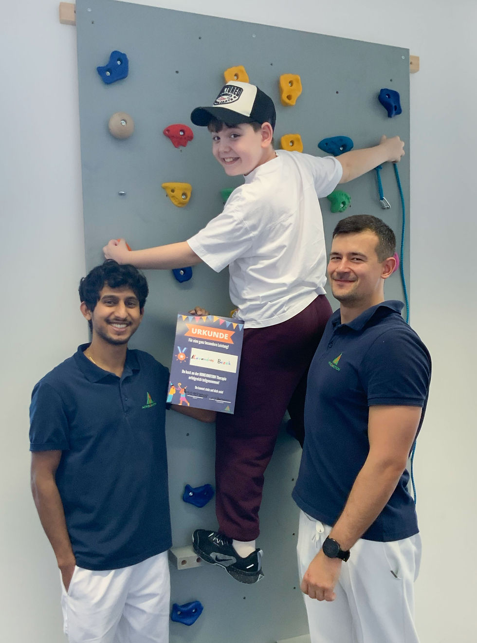 Ein Junge mit einer Kappe klettert eine kleine Kletterwand hinauf und dreht sich dabei lächelnd um. Neben ihm stehen zwei Therapeuten. Alle drei schauen in die Kamera und lächeln.