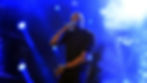 Performer in black shirt raps energetically on stage, holding a mic. Blue lights and smoke create a dramatic concert atmosphere.