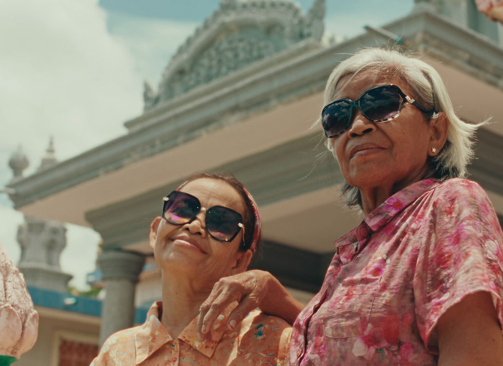 Two elderly women in colorful shirts and sunglasses smile confidently outside an ornate building under a clear sky, exuding joy and warmth.