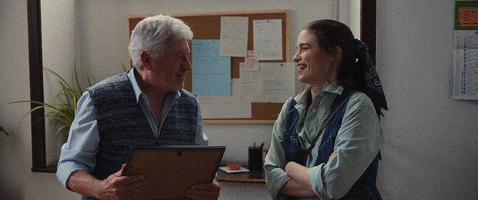 Elderly man and young woman smiling at each other in an office. Man holds a frame; bulletin board and calendar in the background. Relaxed mood.