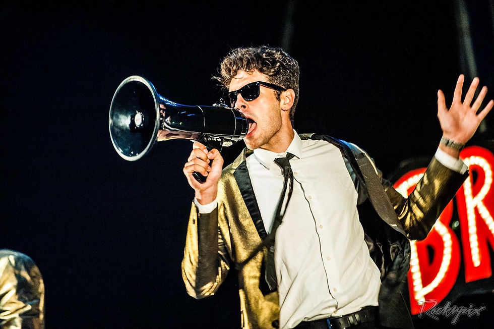 Performer in gold jacket and tie shouting into a megaphone on stage, dark background, wide landscape
