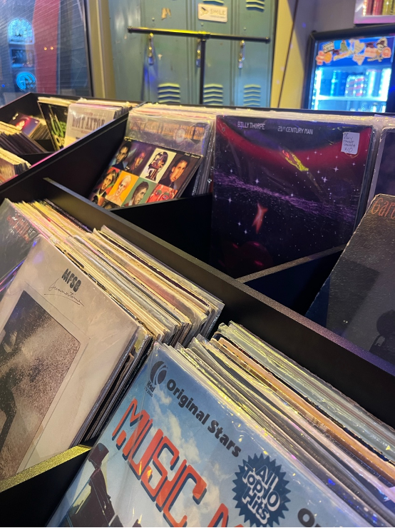 Vinyl records in bins at a store, with retro album covers. Background has blue lockers and a lit vending machine. Warm, nostalgic vibe.