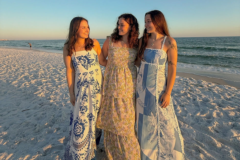 Three women in patterned dresses stand on a sunlit beach, smiling at each other. The ocean and a clear sky form the serene backdrop.