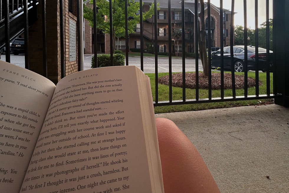 Book titled "Love & Gelato" held by a person sitting on concrete. Background shows a black car, green grass, and a brick building.