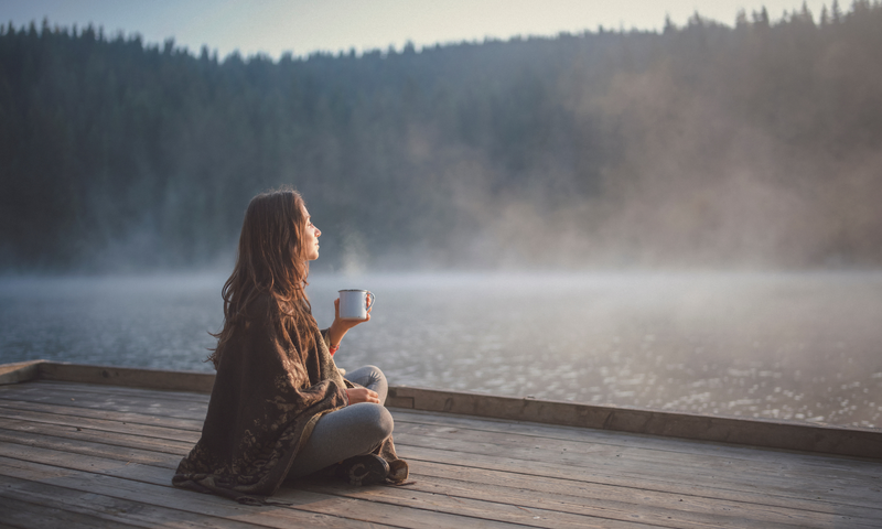 Woman sitting on deck calmly sipping tea overlooking lake