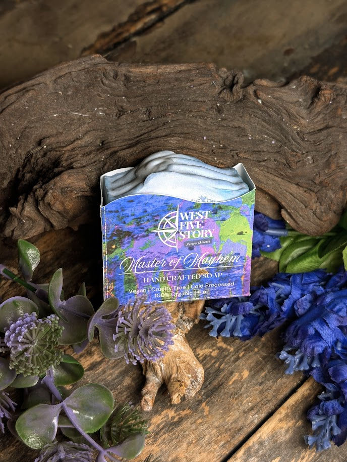 A handcrafted bar of West Five Story “Master of Mayhem” soap sits on a rustic wooden surface beside a piece of driftwood, sur