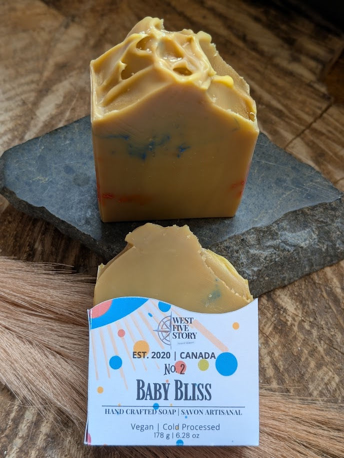 A professional product photograph of "Baby Bliss" Artisan Botanical Soap by West Five Story. The bar features a warm golden-h
