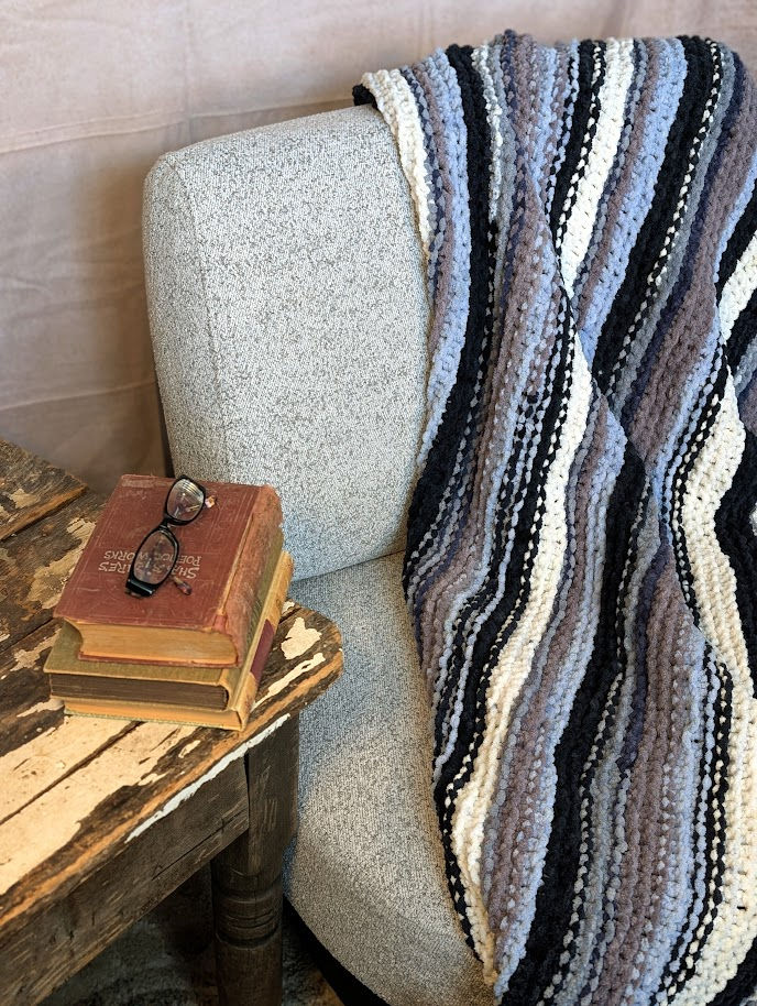 Thumbnail: “A light gray upholstered armchair draped with a plush striped blanket in blue, gray, black, and white, next to a rustic wood