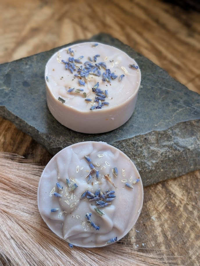 Round Pucks: A lifestyle shot of two 2.5 oz round "Spring Awakening" soap pucks. The soft purple soap is topped with dried la
