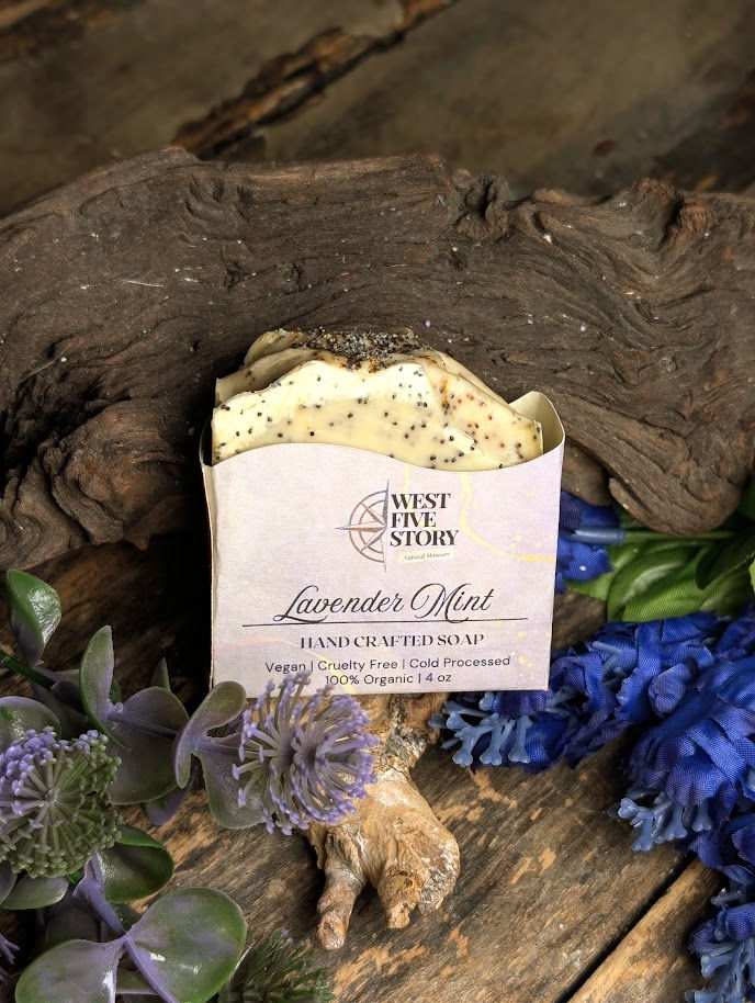A handcrafted bar of West Five Story “Lavender Mint” soap rests on a rustic wooden surface beside a piece of driftwood, surro