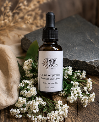 A product photo for West Five Story's 'Calm Complexion' Calming Facial Serum in an amber glass dropper bottle, resting on a wooden surface surrounded by white flowers, a dark slate piece, and a soft cloth