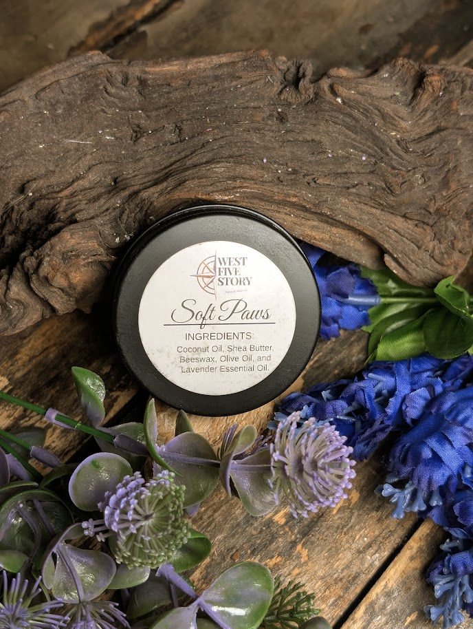 A handcrafted tin of West Five Story “Soft Paws” balm sits on a rustic wooden surface beside a piece of driftwood, surrounded