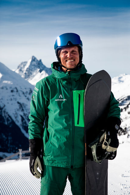 Snowboarder lächelt mit Board vor schneebedeckten Bergen, trägt Helm und Jacke.